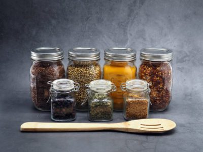 A collection of spices in mason jars, behind a wooden spoon. Product photography staged on a grey backdrop.